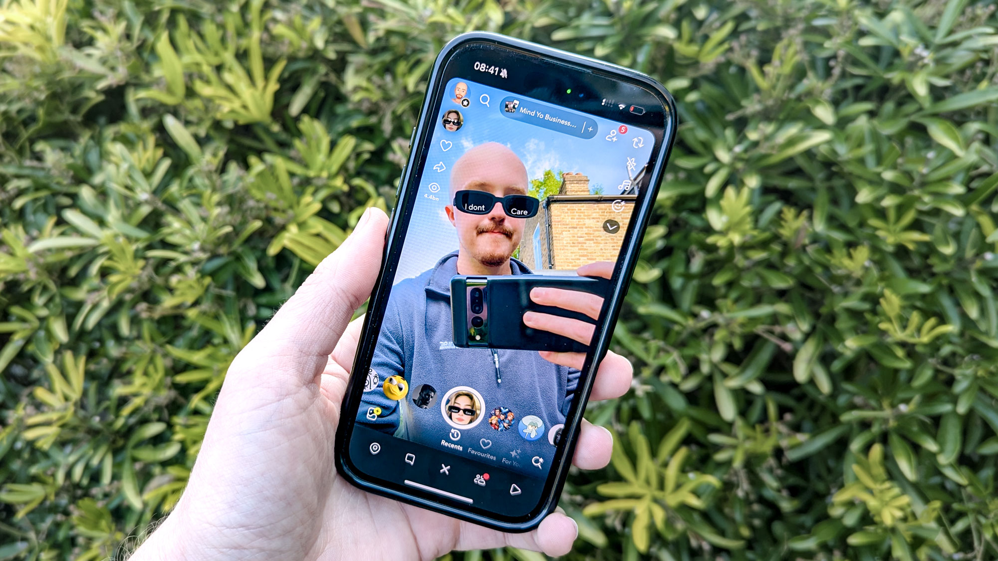 hand holding iPhone with Snapchat camera filter showing person with glasses on screen with bush in background