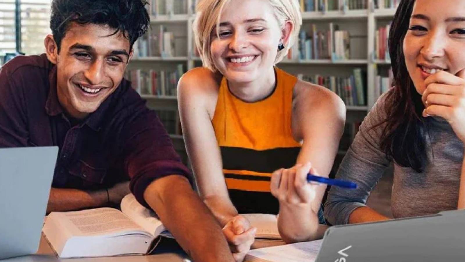 3 students (1 boy and 2 girls) sitting in a library with books and laptops. They're smiling.