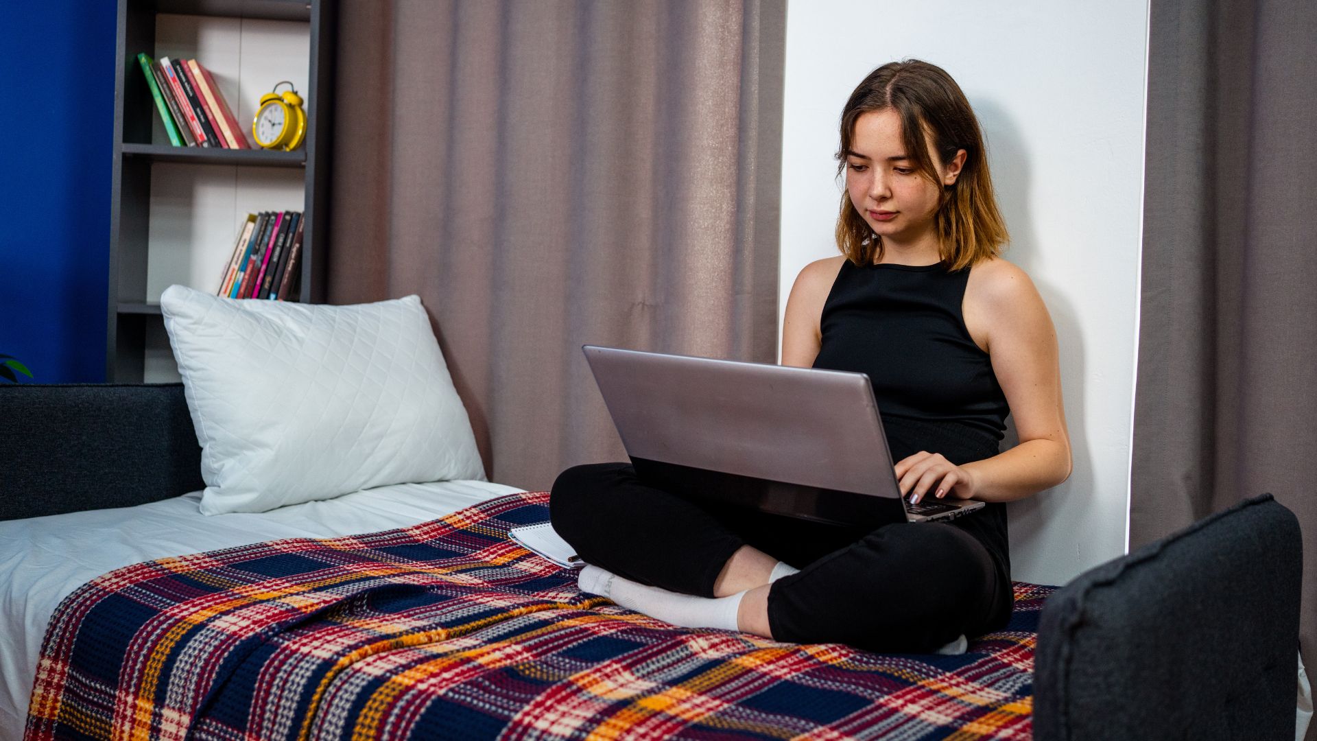 A young female college student with shoulder length brown hair looks at her laptop while sitting on her dorm bed.