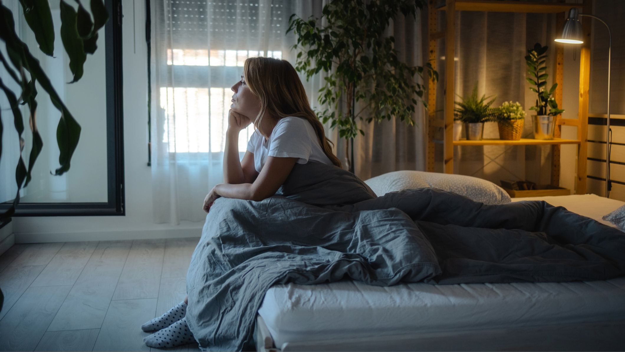 A woman sitting on the edge of her mattress resting her head on her hand and thinking