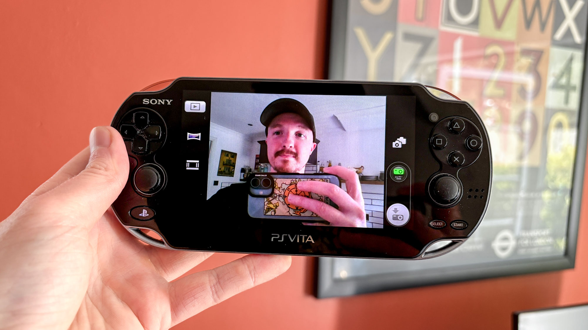 PS Vita في اليد يعرض الكاميرا على شاشة لشخص يحمل iPhone