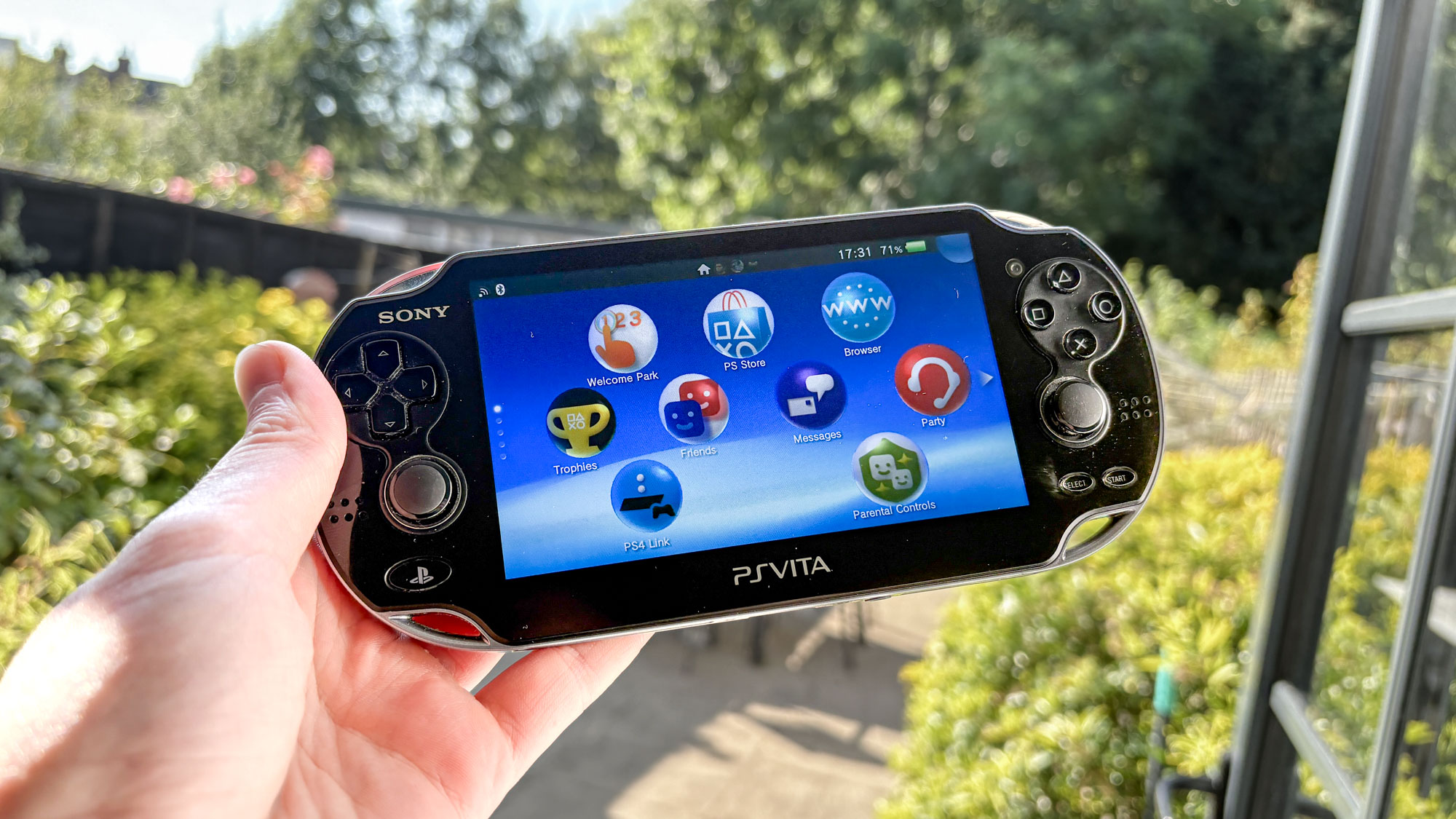 PS Vita في اليد مع حديقة خارجية في الخلفية مع قائمة الشاشة الرئيسية معروضة