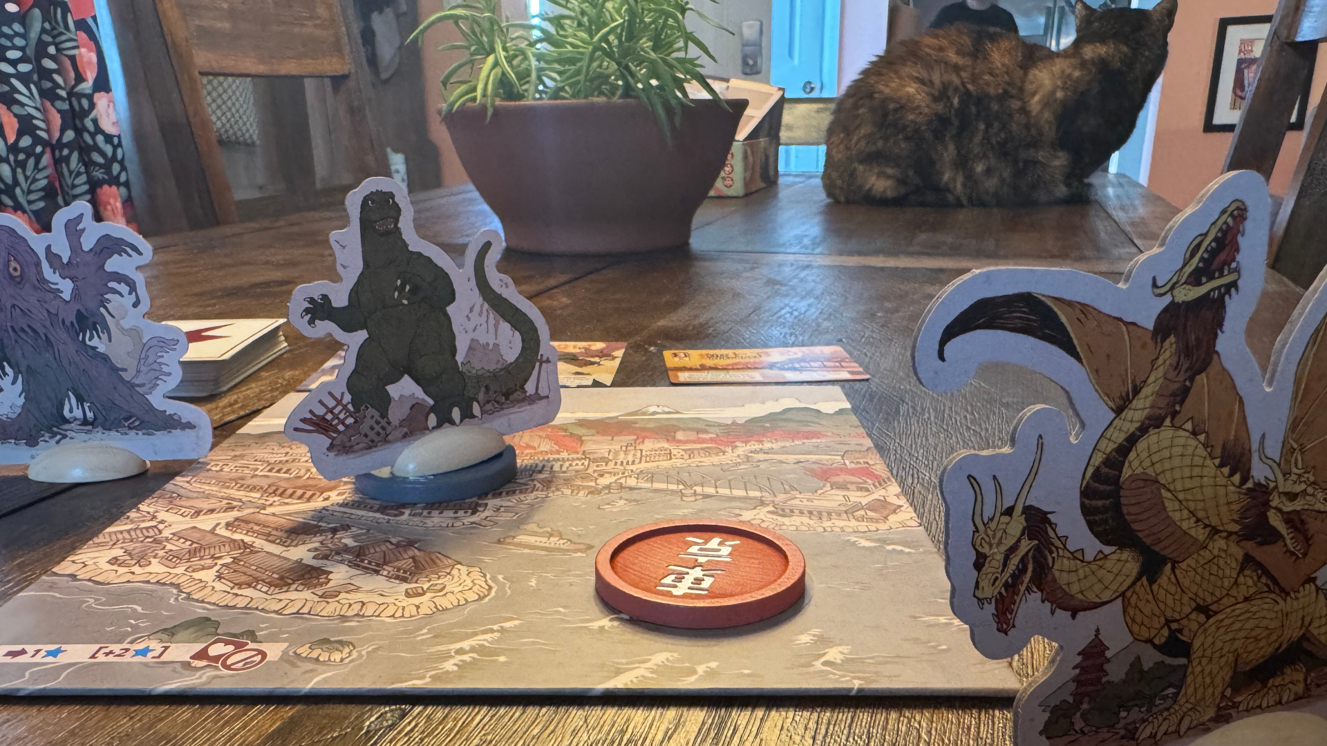 رموز وبطاقات متعددة للعبة King of Tokyo: Godzilla على سطح خشبي، مع قطة ظاهرة في الخلفية
