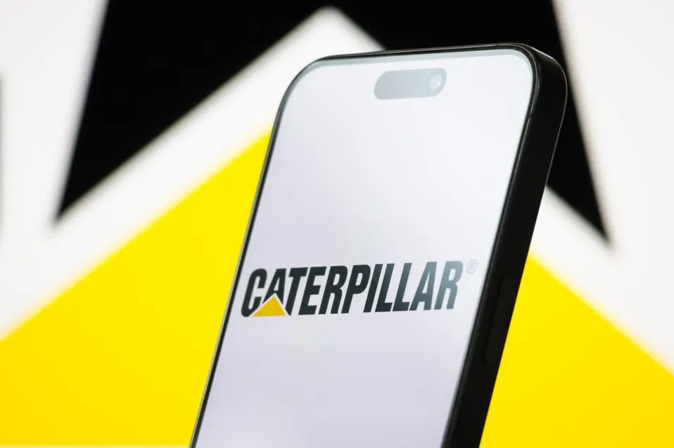 تشونغتشينغ، الصين - 26 أبريل: في هذا الرسم التوضيحي للصور، يتم عرض شعار شركة Caterpillar Inc. ... أكثر على شاشة هاتف ذكي، مع العلامة التجارية الصفراء والسوداء المميزة للشركة المرئية في الخلفية، في 26 أبريل 2025، في تشونغتشينغ، الصين. (صورة توضيحية لـ Cheng Xin/Getty Images)