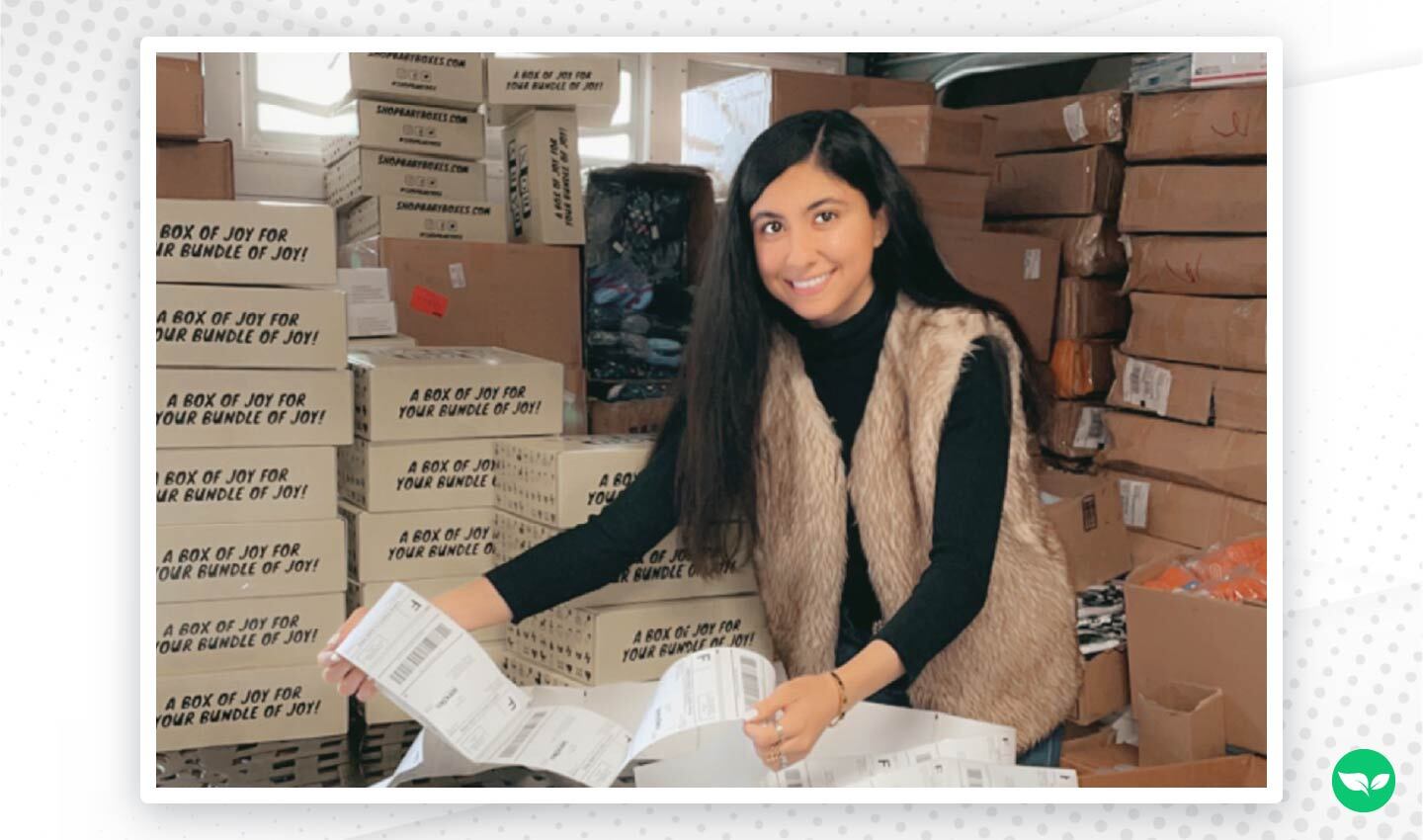 Zarina Bahadur sorride mentre conta le etichette di spedizione in un magazzino pieno di pacchi impilati di 123 Baby Box.