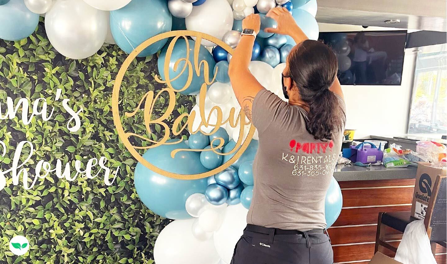 Karen Kaplan sta assemblando palloncini blu e bianchi per un baby shower, sistemando le decorazioni su uno sfondo verde lussureggiante.