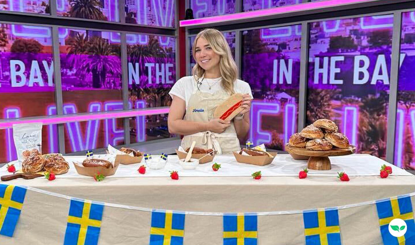 Amelia Udaily en el plató de Live in the Bay, de pie detrás de una mesa decorada con pasteles suecos y perritos calientes.