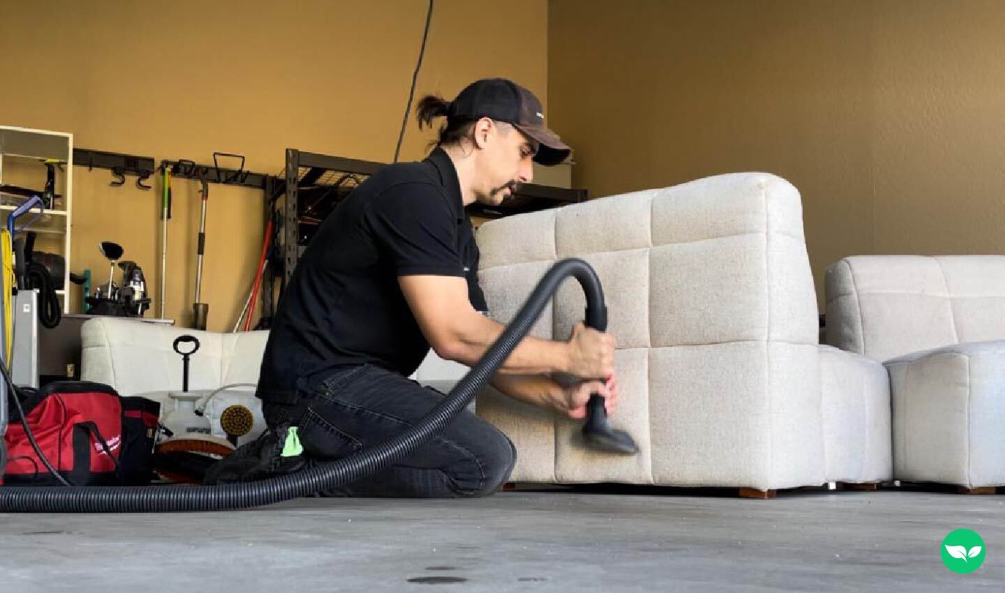 Kyle Strood è inginocchiato in un garage, intento a pulire un divano beige con il tubo di un aspirapolvere professionale, circondato da attrezzature e strumenti di lavoro.