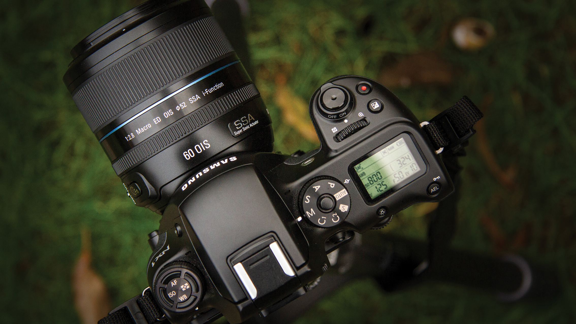 كاميرا Samsung DSLR سوداء مع عدسة ماكرو 60 مم مثبتة، تظهر عناصر التحكم العلوية والشاشة، موضوعة على خلفية عشبية خضراء ضبابية