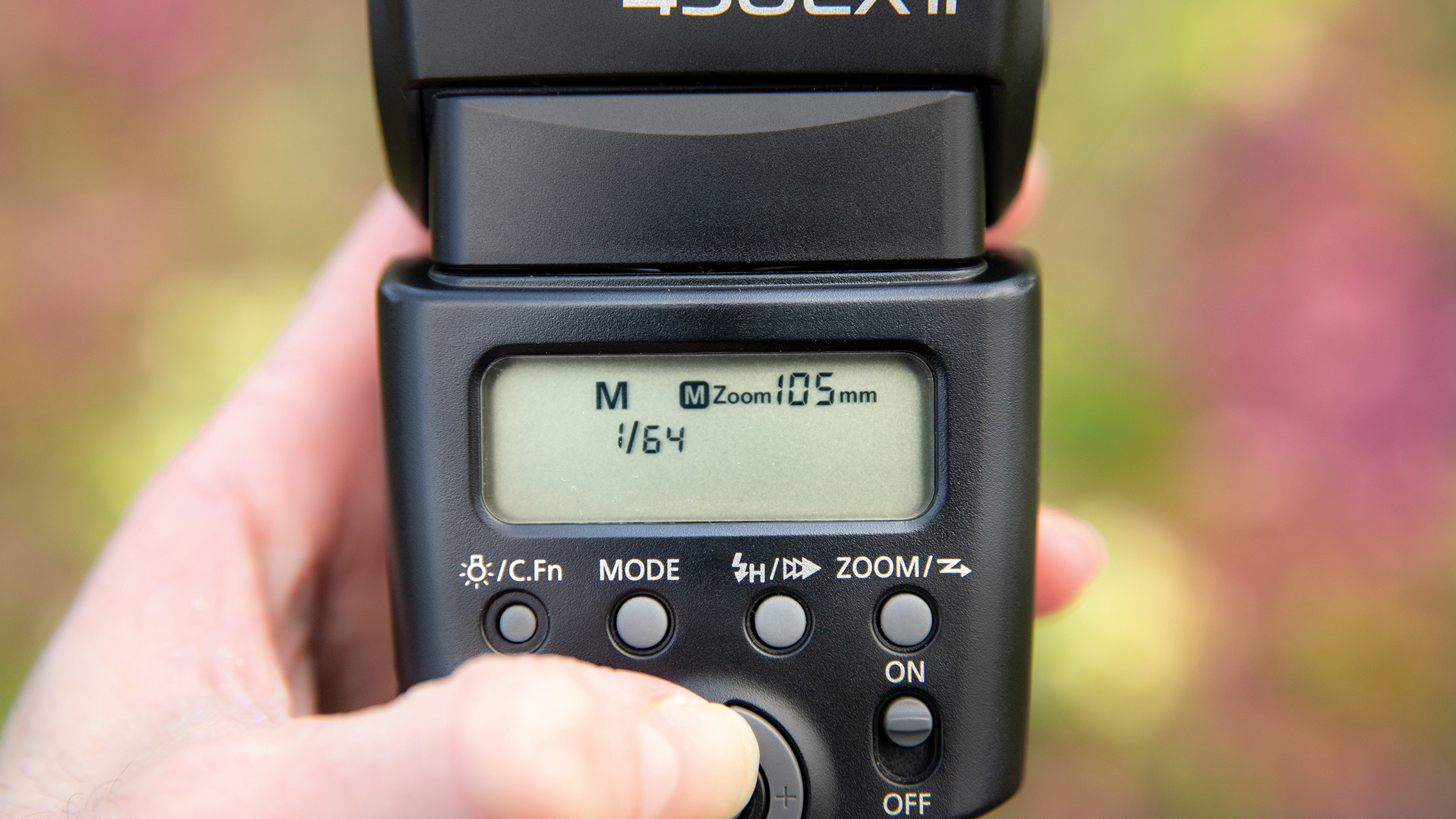 لقطة مقربة ليد تضبط الإعدادات على وحدة فلاش الكاميرا. تعرض الشاشة "M Zoom 105mm 1/64" مع أزرار للوضع والتقريب والطاقة. خلفية زهرية ضبابية.