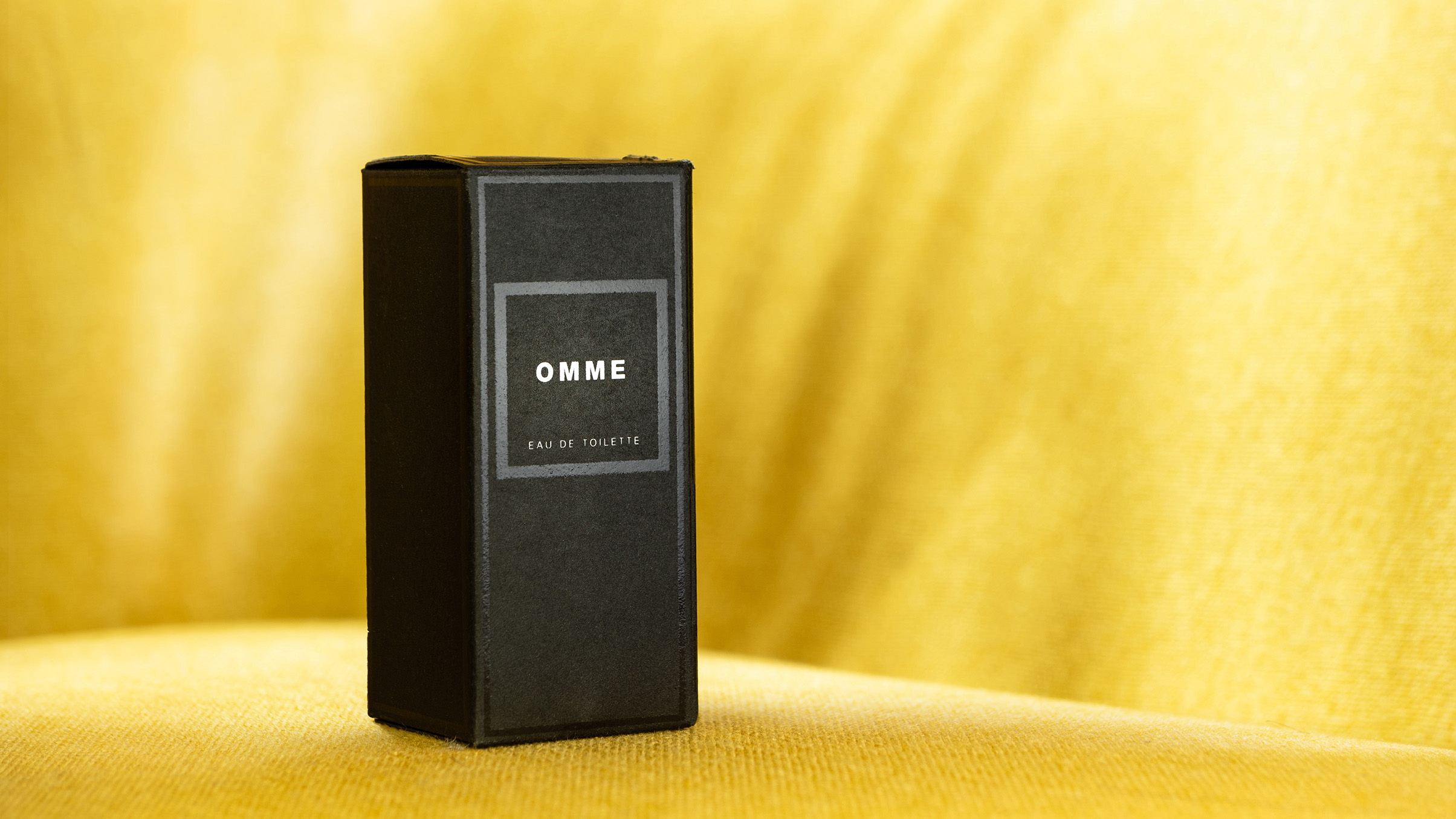صندوق أسود أنيق مكتوب عليه "OMME Eau de Toilette" موضوع على خلفية صفراء ناعمة
