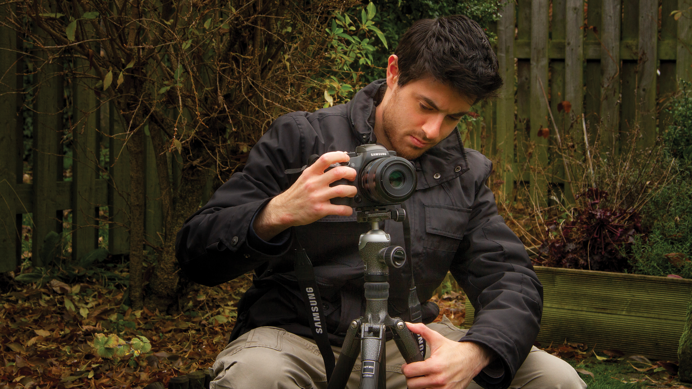 رجل يضبط كاميرا DSLR على حامل ثلاثي في حديقة بها أوراق جافة وخلفية سياج خشبي