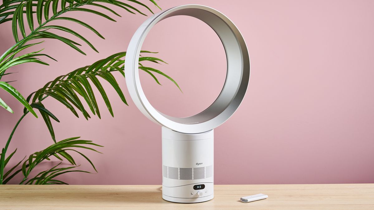 مروحة Dyson Cool CF1 موضوعة بزاوية طفيفة نحو اليمين على سطح طاولة، أمام نبتة خضراء مورقة وجدار وردي. جهاز التحكم عن بعد موضوع على الطاولة إلى يمين المروحة.