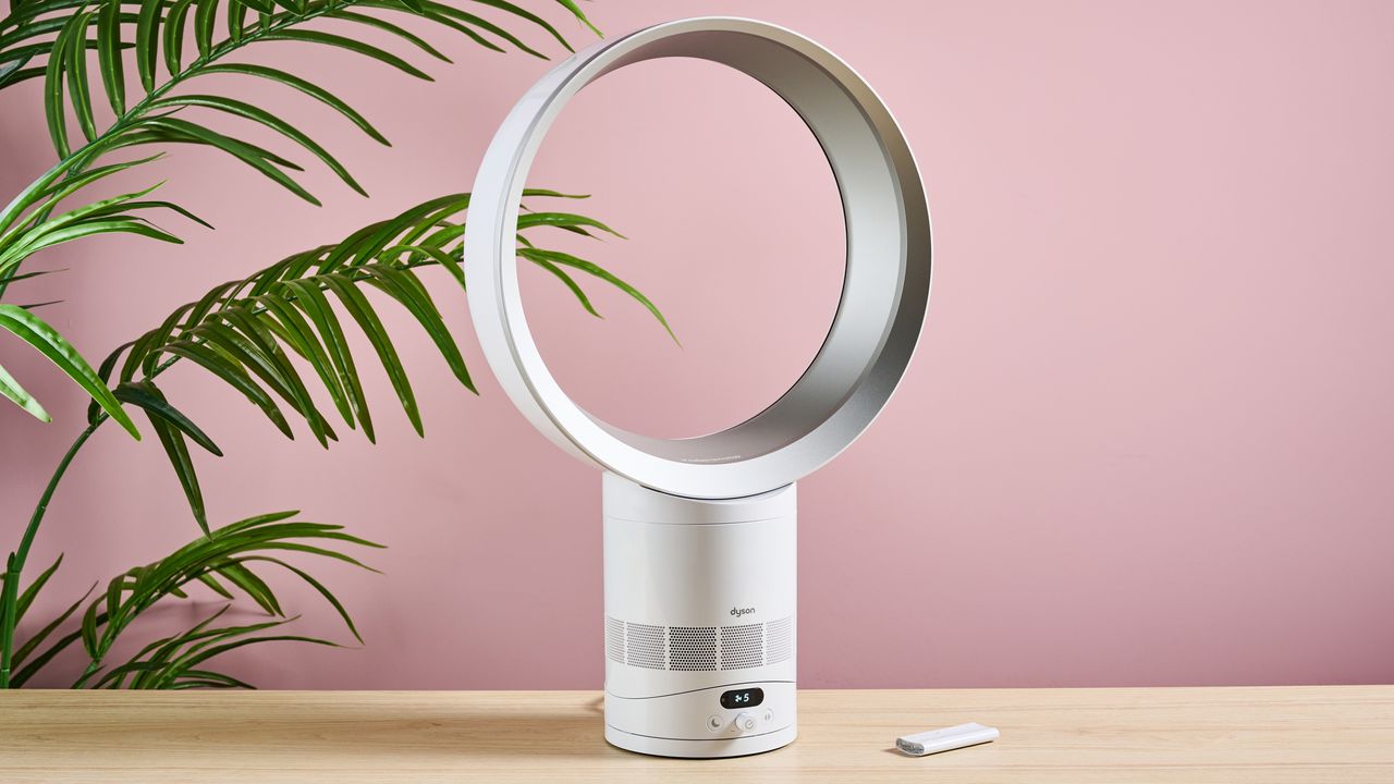 مروحة Dyson Cool CF1 موجهة قليلاً إلى اليمين على سطح طاولة أمام نبات أخضر مورق وجدار وردي. جهاز التحكم عن بعد موجود على الطاولة إلى يمين المروحة.