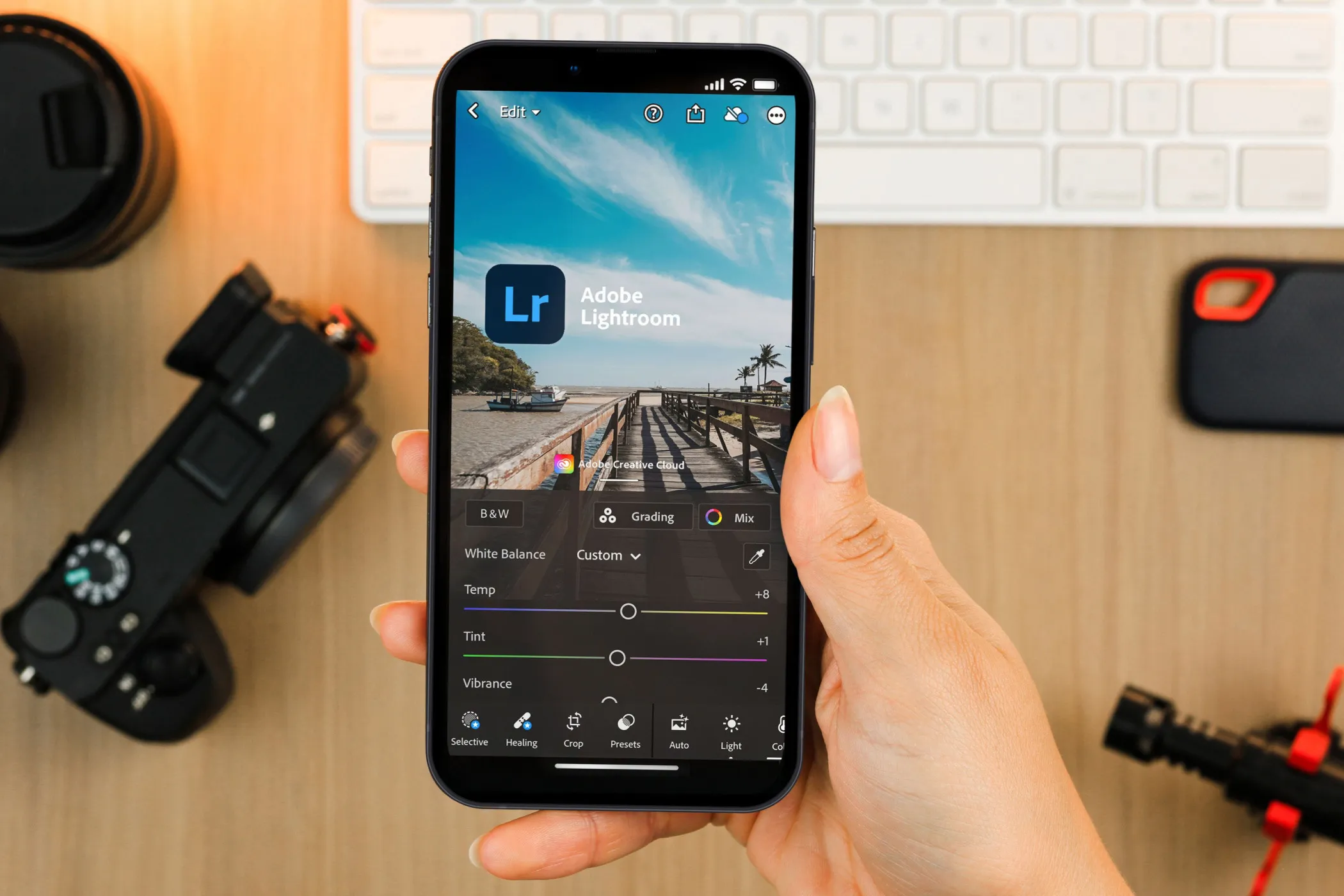 Hand holding smartphone, displaying Lightroom Mobile app, with camera equipment in the background
