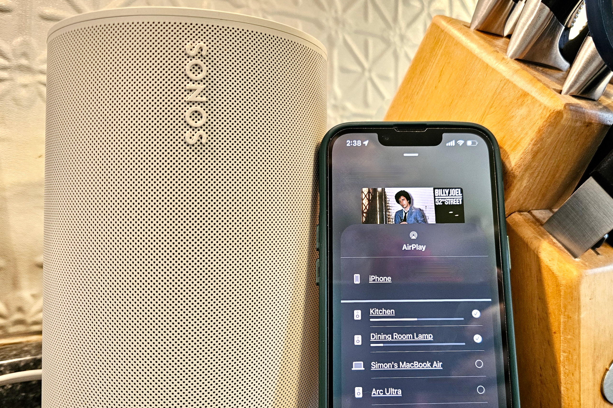 A Sonos Era 100 speaker next to an iPhone showing AirPlay speaker options.