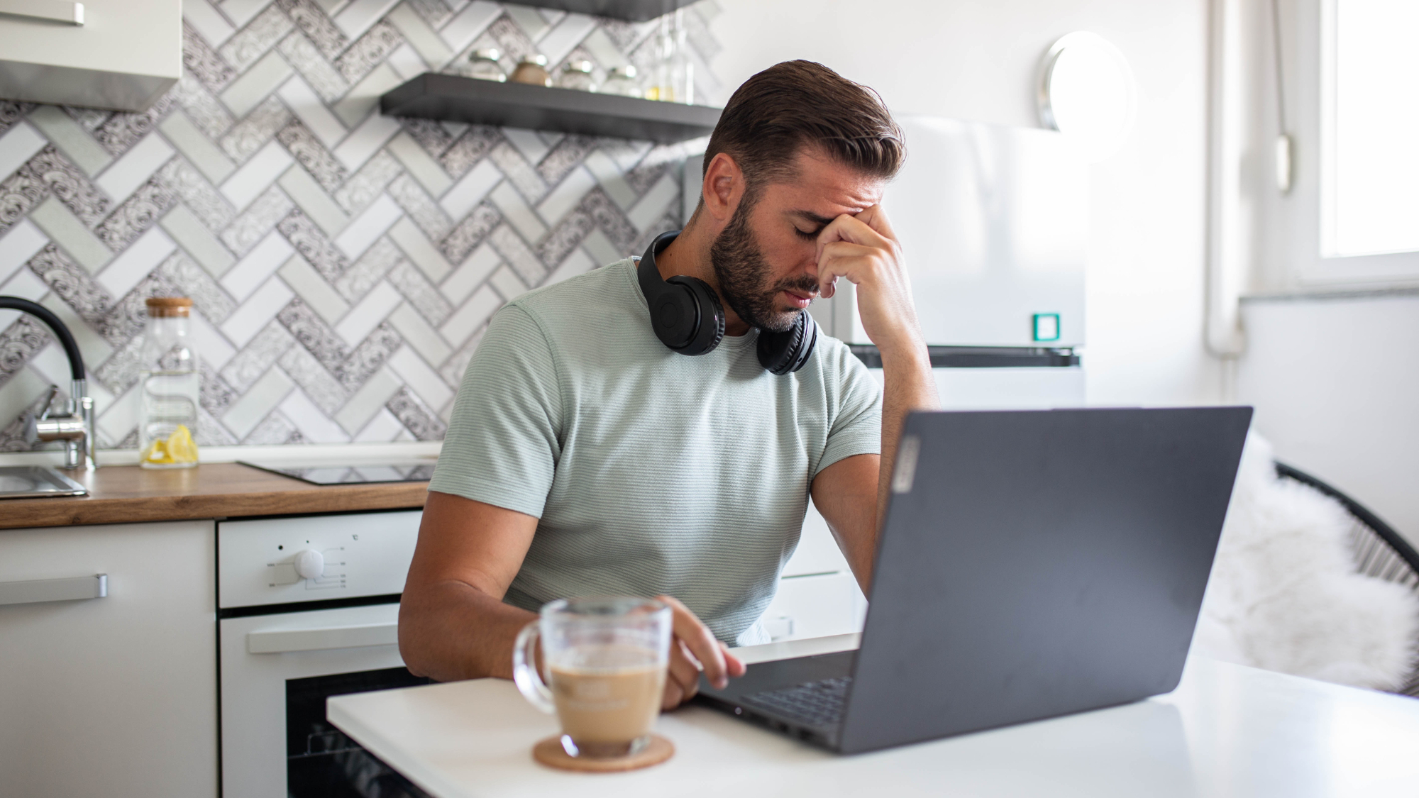 Stressed man in a kitchen sat in front of a laptop with a coffee