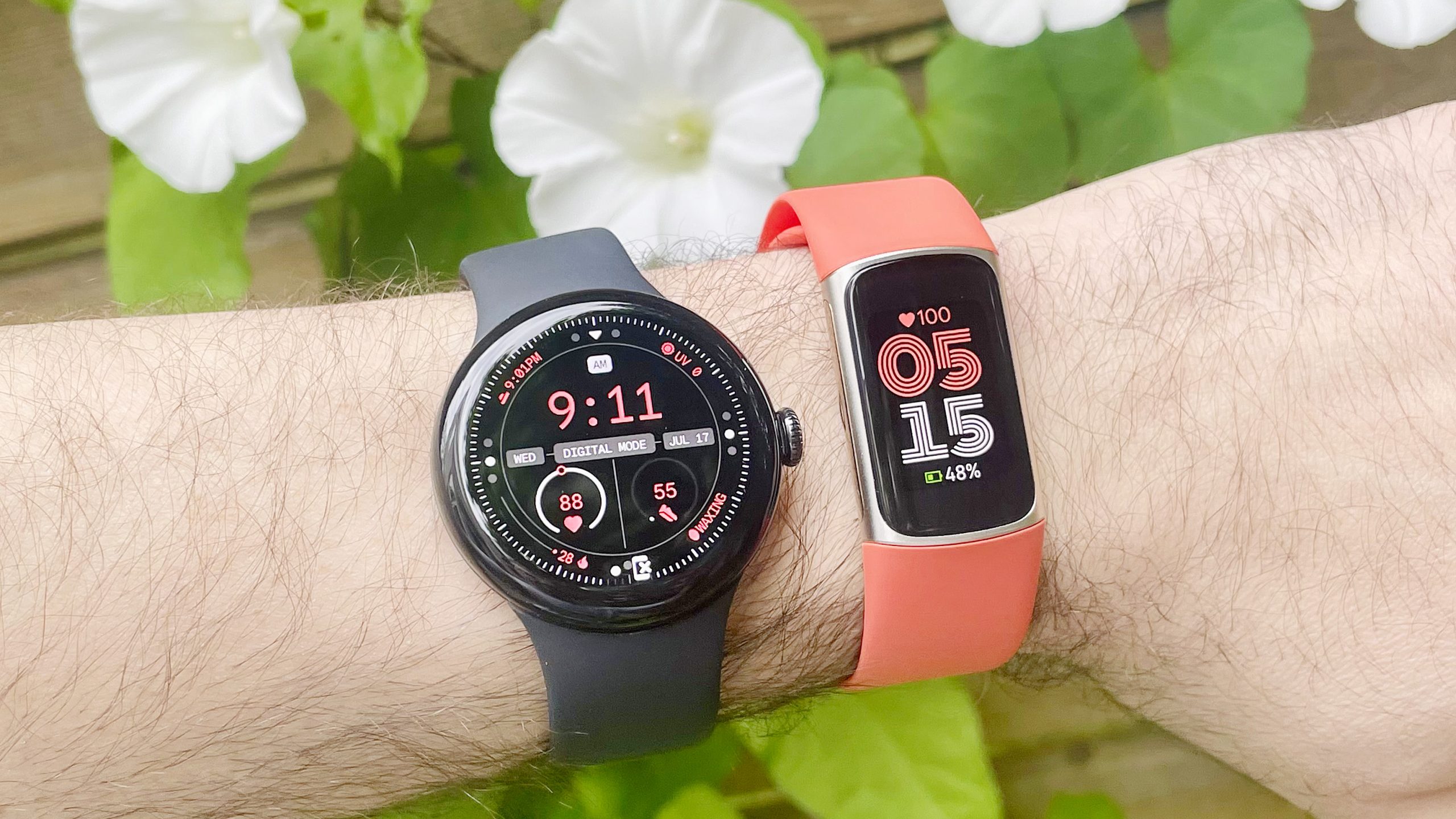 The Pixel Watch 3 on a black strap next to the Fitbit Charge 6 on an orange strap on a user's wrist with white flowers in the background