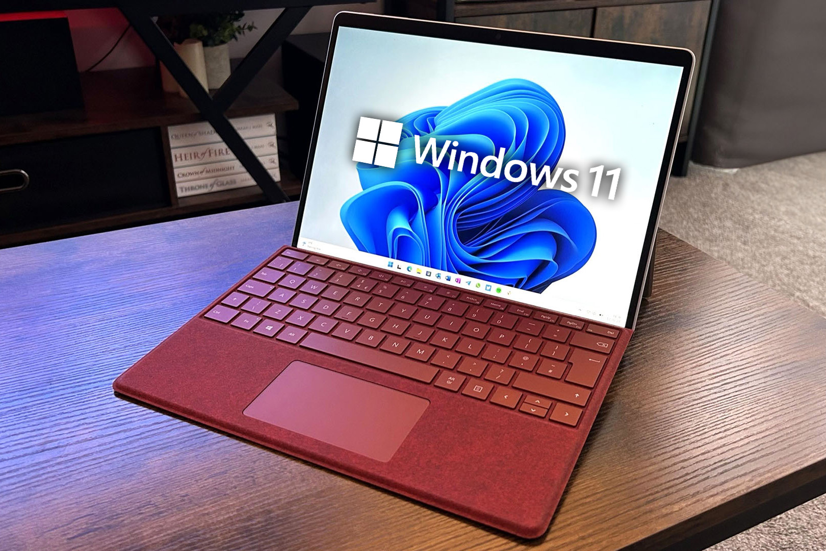 شعار Windows 11 مُركّب على شاشة جهاز Microsoft Surface Pro 9 مع لوحة مفاتيح حمراء موضوعة على مكتب خشبي