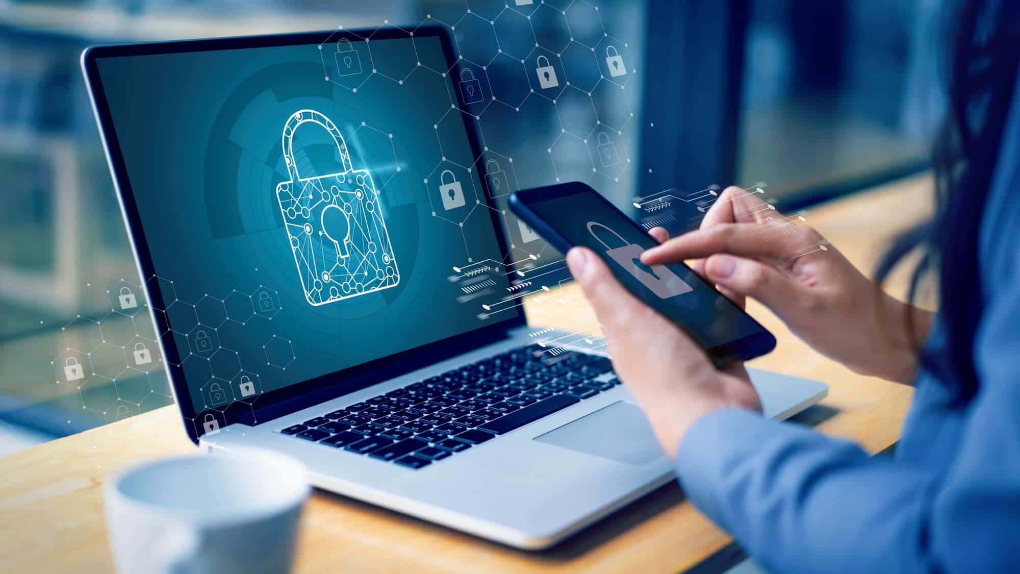 صورة لأيدي امرأة تحمل هاتفًا ذكيًا عليه رمز قفل، أمام جهاز كمبيوتر محمول عليه أيضًا رمز قفل.