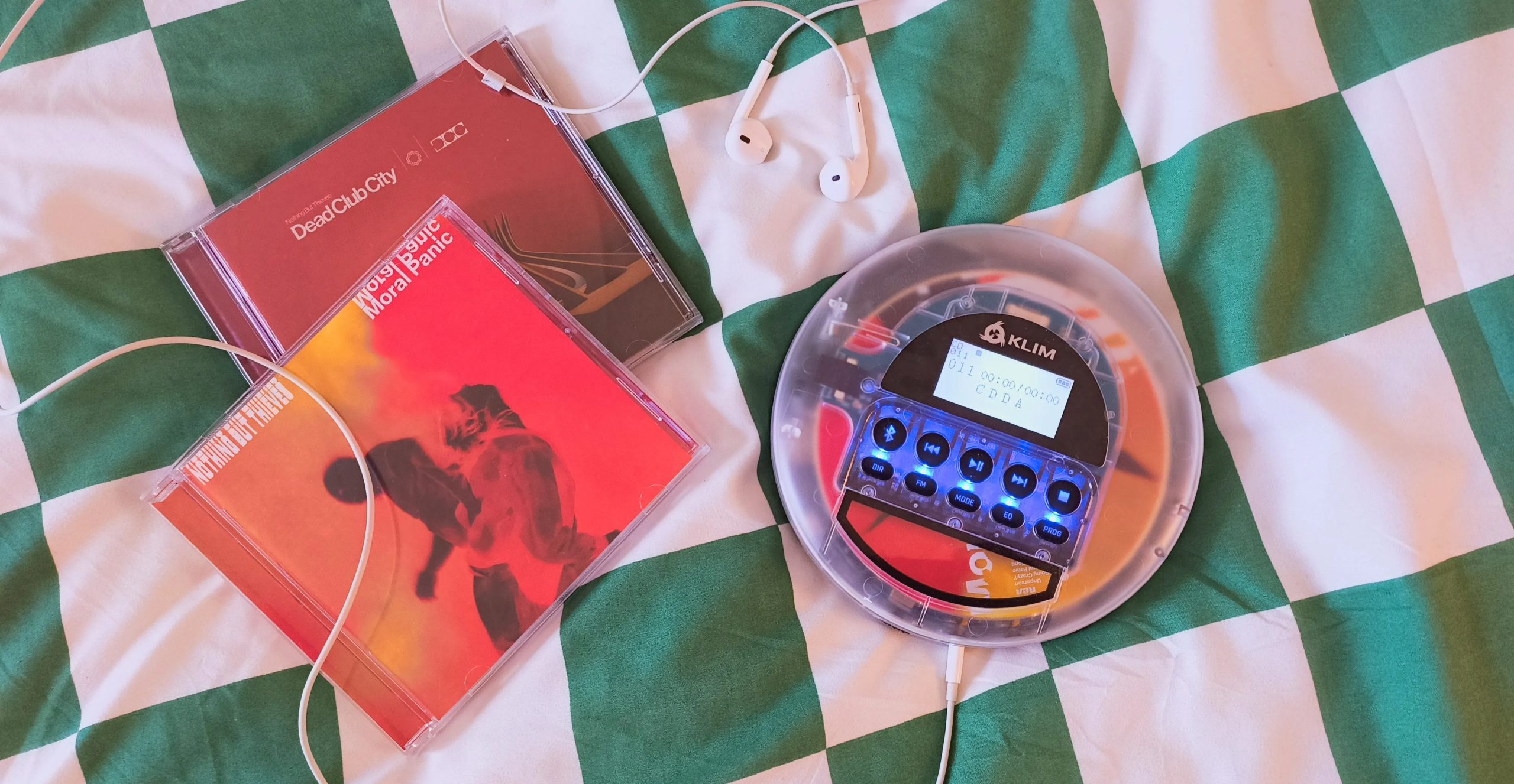 A Klim Nomad CD player and headphones on a bed next to two Nothing But Thieves CDs