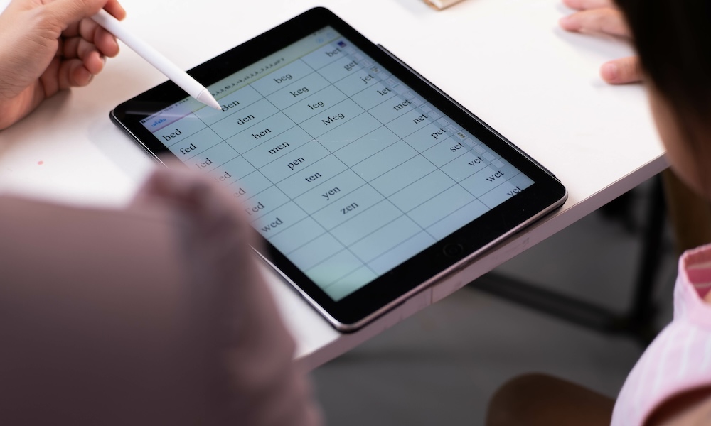 طفلة آسيوية سعيدة تتحدث مع والدتها باستخدام جهاز لوحي رقمي لأداء الواجبات المدرسية أو الدراسة عبر الإنترنت. فتاة صغيرة سعيدة وبالغة جالستان على طاولة يتعلمان معًا على جهاز iPad. العودة إلى المدرسة.