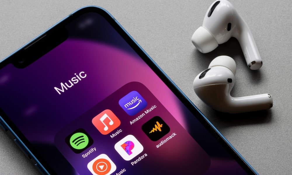 صورة مقربة لهاتف iPhone يعرض تطبيقات بث الموسيقى المختلفة مثل Spotify و Apple Music و Amazon Music و YouTube Music و Pandora و Audiomack، بجانب زوج من سماعات AirPods Pro.