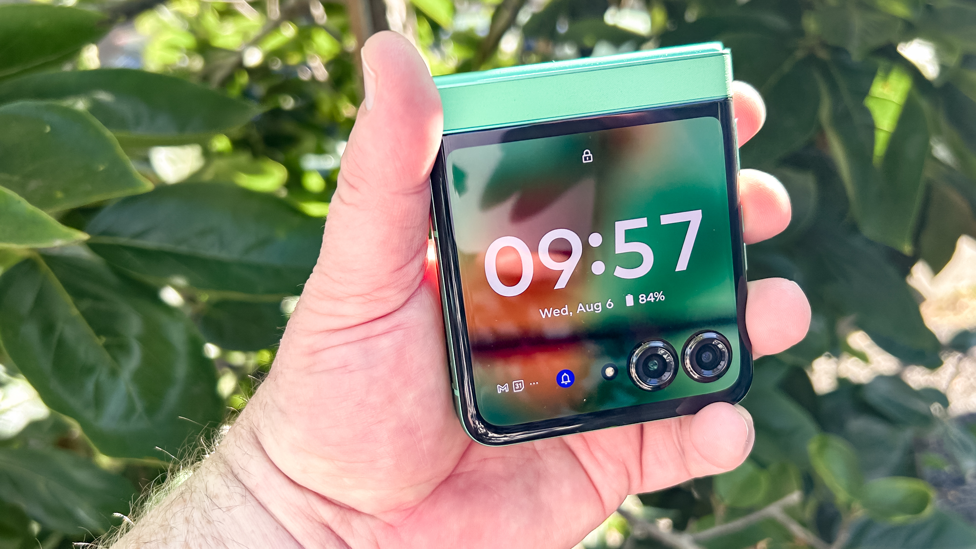 شخص يحمل هاتف Motorola Razr 2025 مطويًا أمام شجيرة خضراء كبيرة، تظهر الشاشة الخارجية مضاءة بالتاريخ والوقت ومستوى البطارية