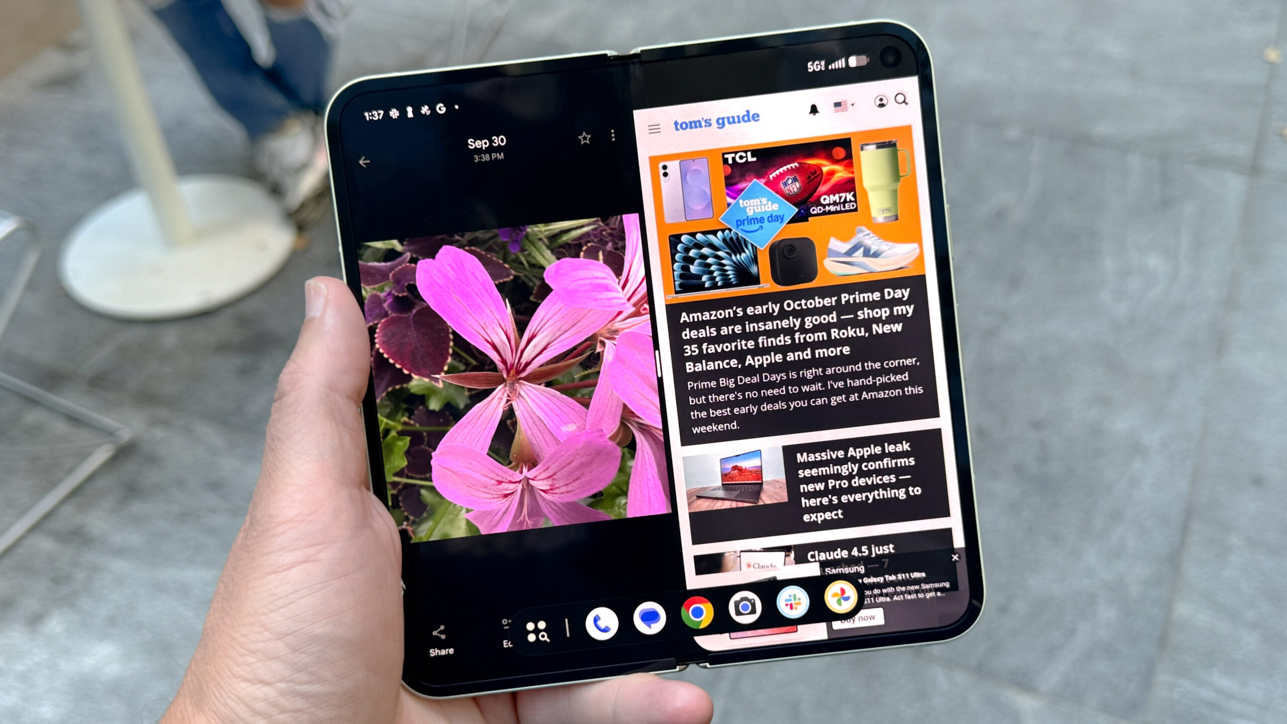 شخص يحمل هاتف Pixel 10 Pro Fold مفتوحًا بالكامل، يعرض صورة زهرة على جانب وصفحة تومز جايد الرئيسية على الجانب الآخر، مع شريط التطبيقات في أسفل الشاشة في منطقة حضرية
