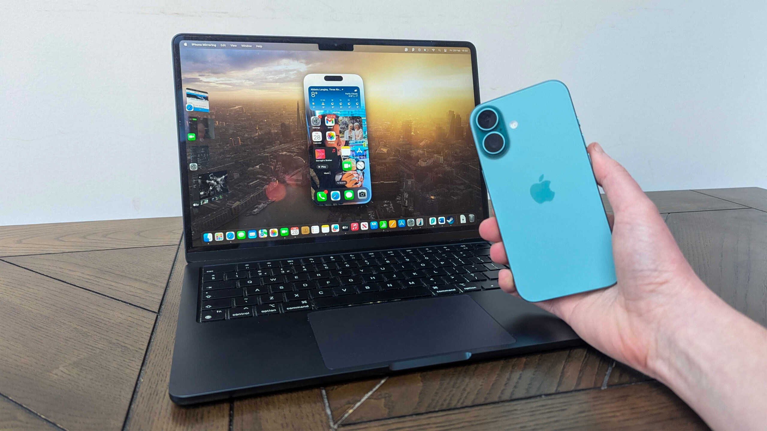 هاتف iPhone 16 في اليد أمام جهاز MacBook Air 13-inch M3 على الطاولة