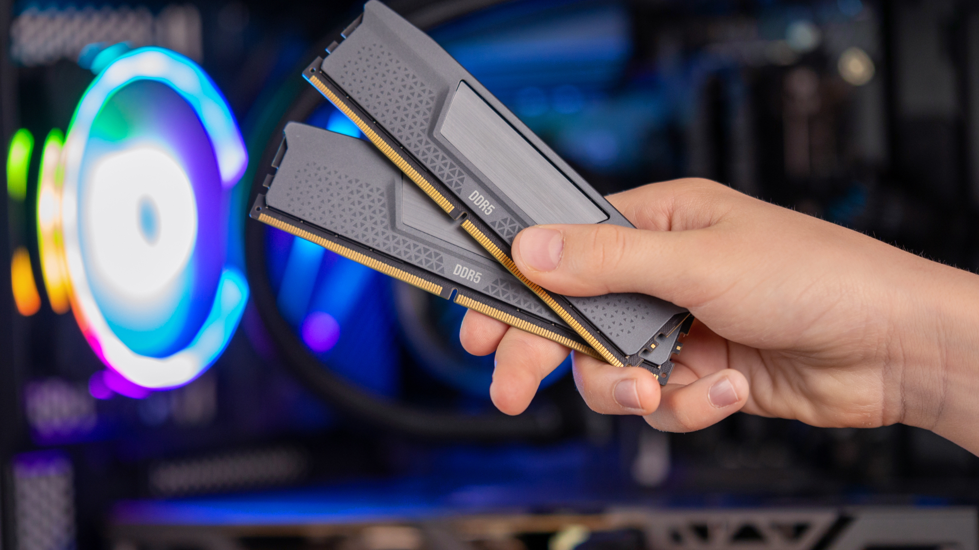 يد تحمل وحدتي ذاكرة DDR5 RAM أمام جهاز كمبيوتر ألعاب بإضاءة RGB