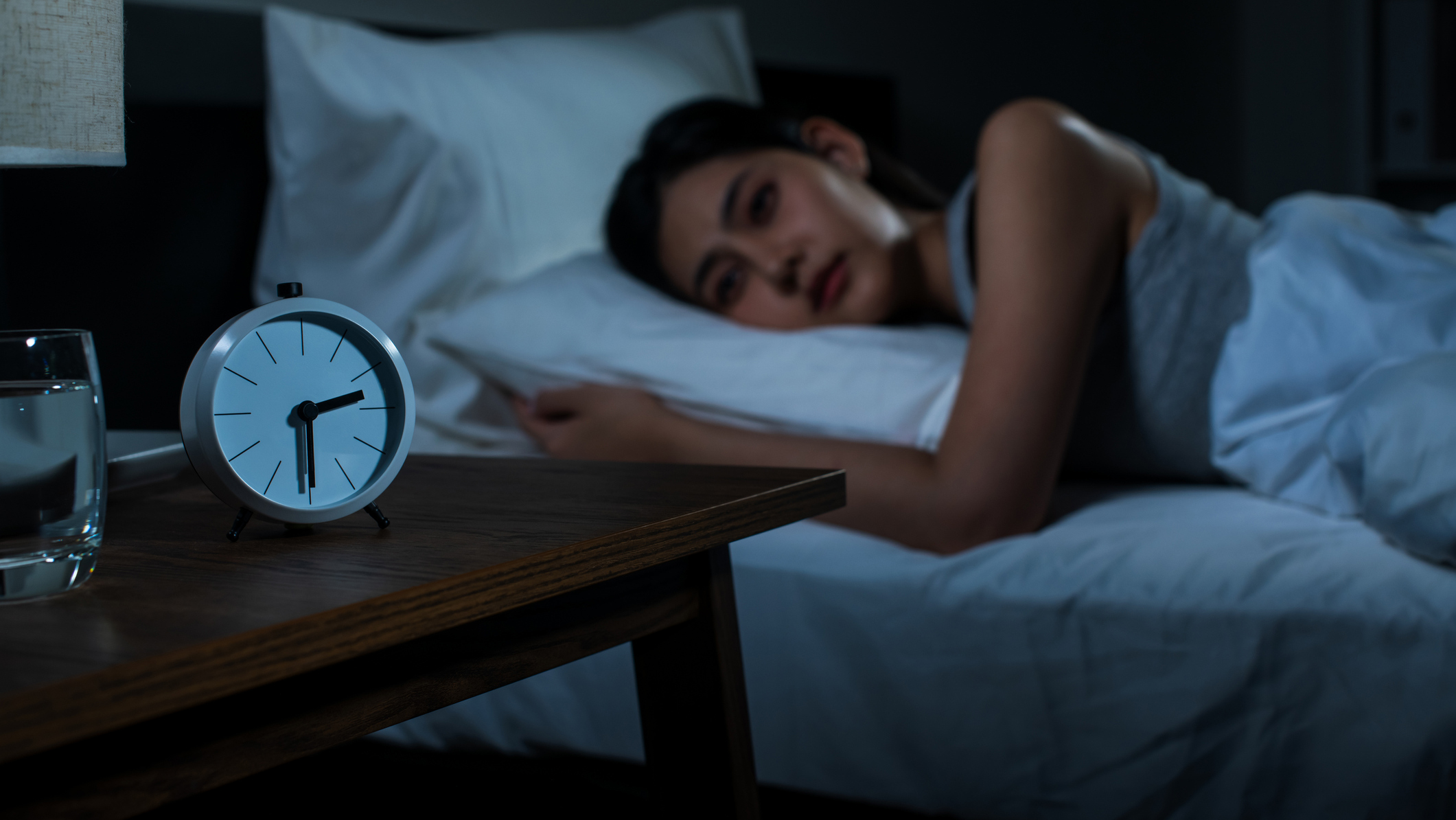 A woman with dark brown hair lies on her side staring at the back of a white alarm clock that shows the time as 2.30am