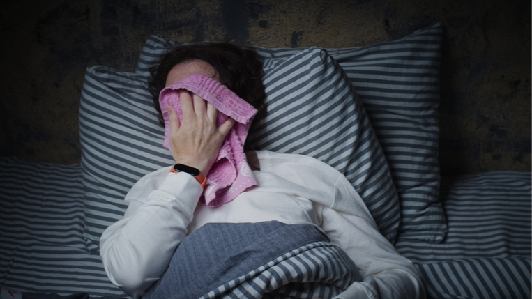 A woman in bed with navy and white stripey sheets holding a pink flannel over her face to cool down, wearing white pyjamas and a red smart watch