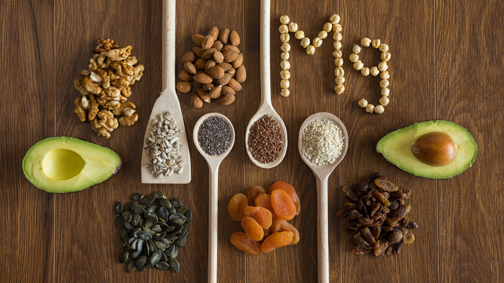 An image showing different magnesium rich foods, including avocados, almonds, black beans, and chia seeds, displayed on wooden spoons placed on a wooden table