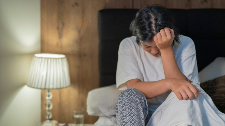 امرأة ترتدي قميصًا أبيض وبنطلون بيجامة تجلس في السرير ورأسها بين يديها.