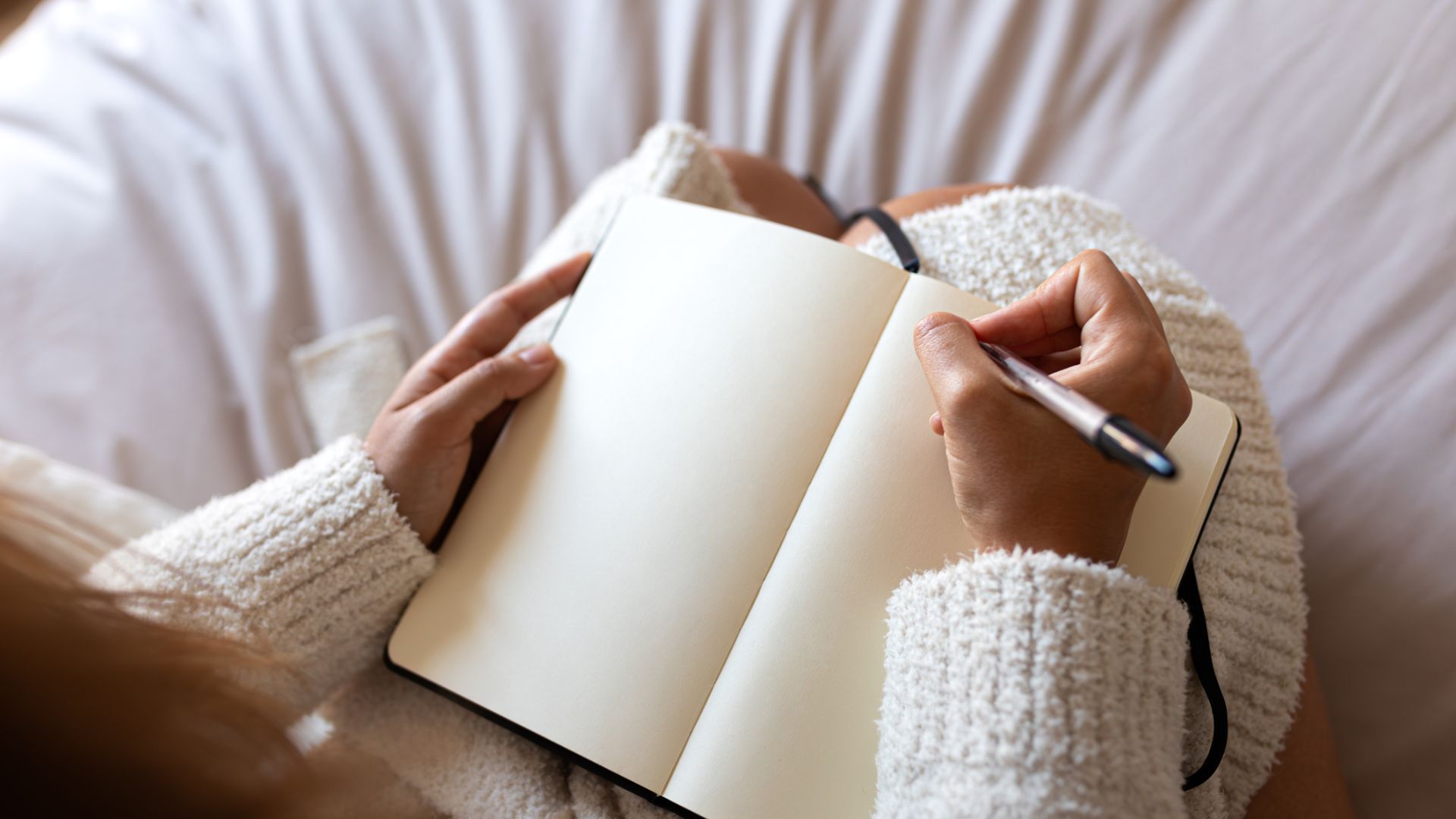 Dos manos de una mujer sosteniendo un cuaderno y un bolígrafo, a punto de empezar a escribir en un diario mientras está en la cama.