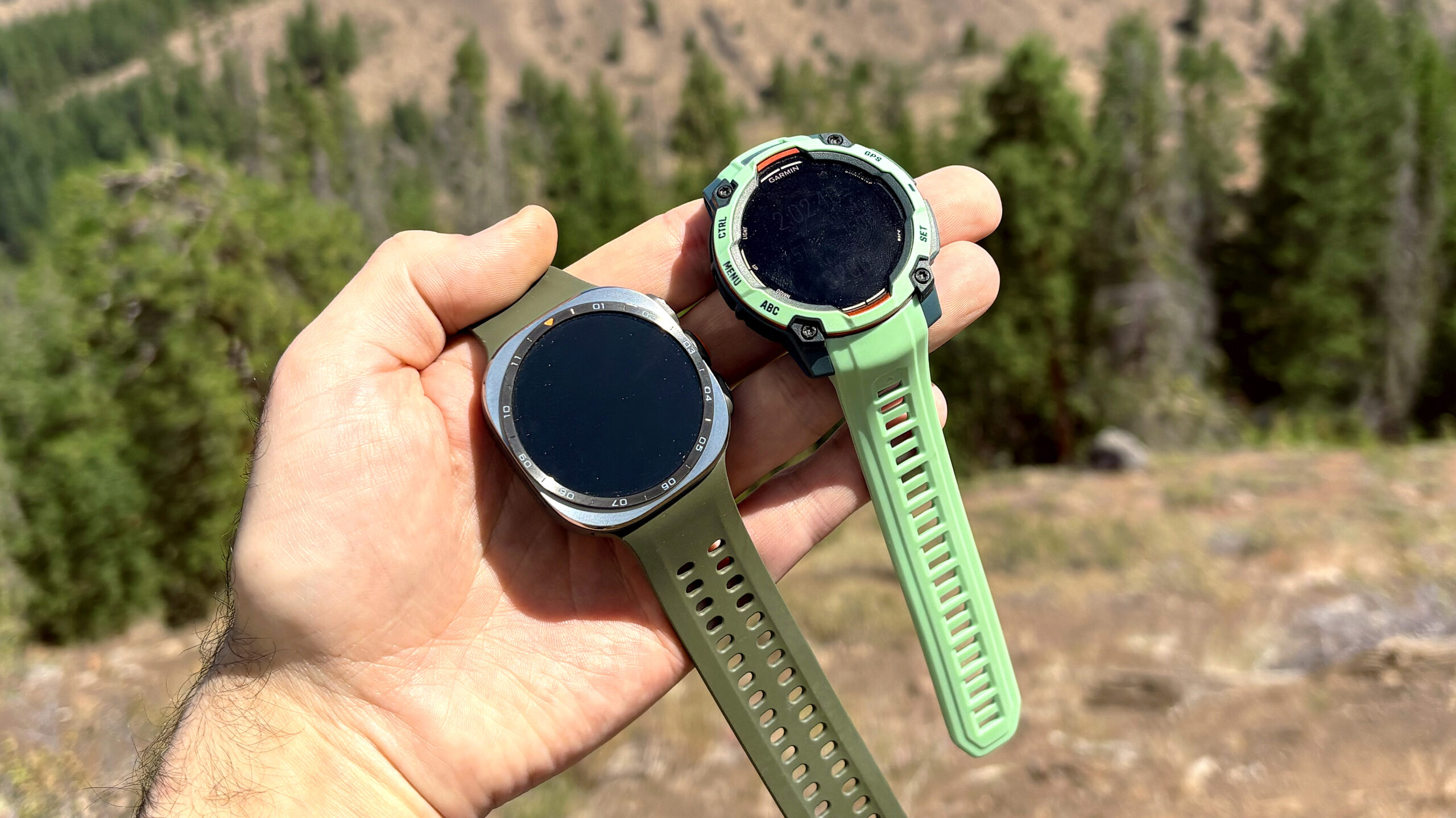لقطة مقربة لساعتي Samsung Galaxy Watch Ultra 2 و Garmin Instinct 3 في الجبال أثناء رحلة ركوب دراجات كهربائية