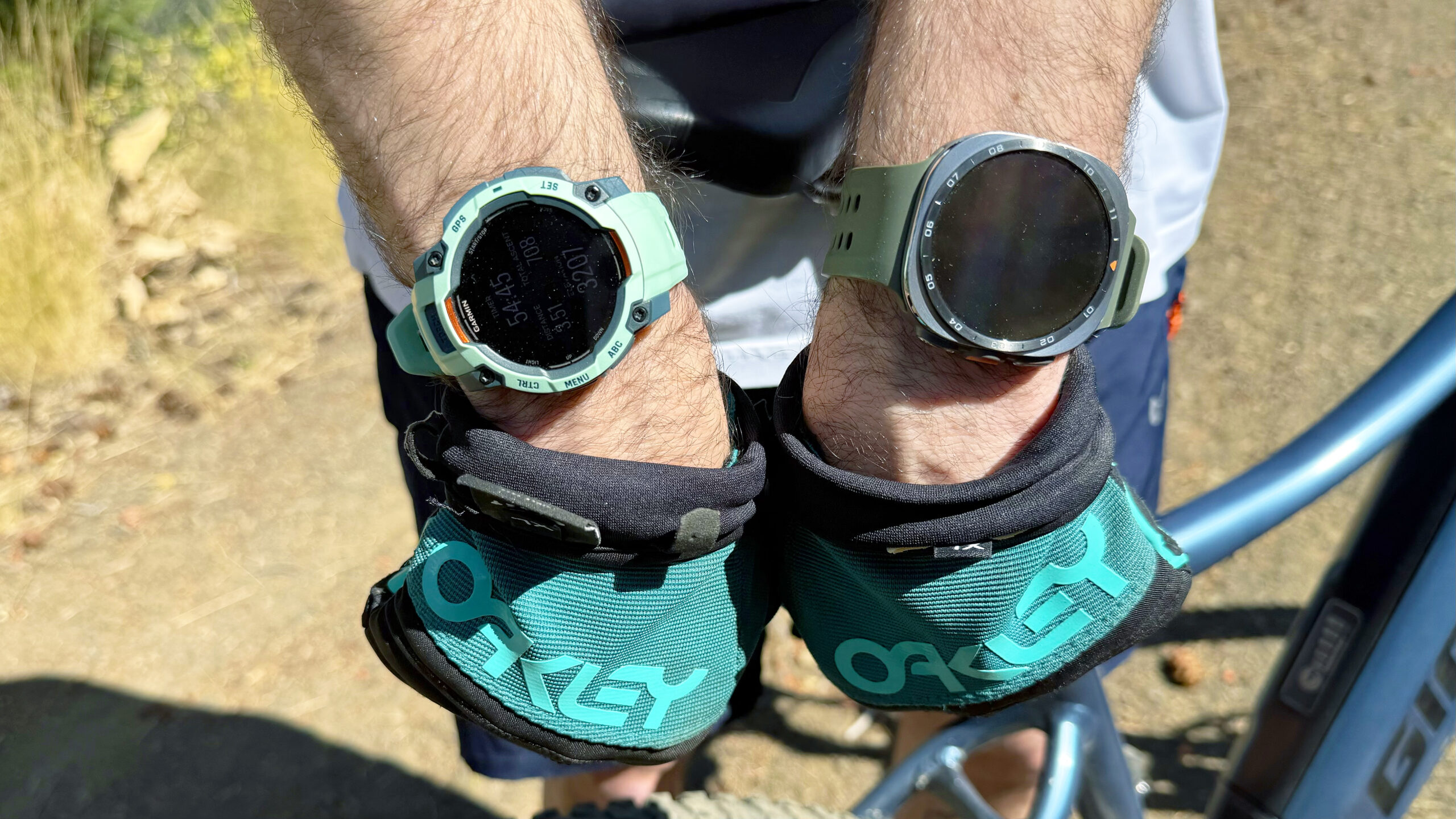 لقطة مقربة لساعتي Samsung Galaxy Watch Ultra 2 و Garmin Instinct 3 في الجبال أثناء رحلة ركوب دراجات كهربائية