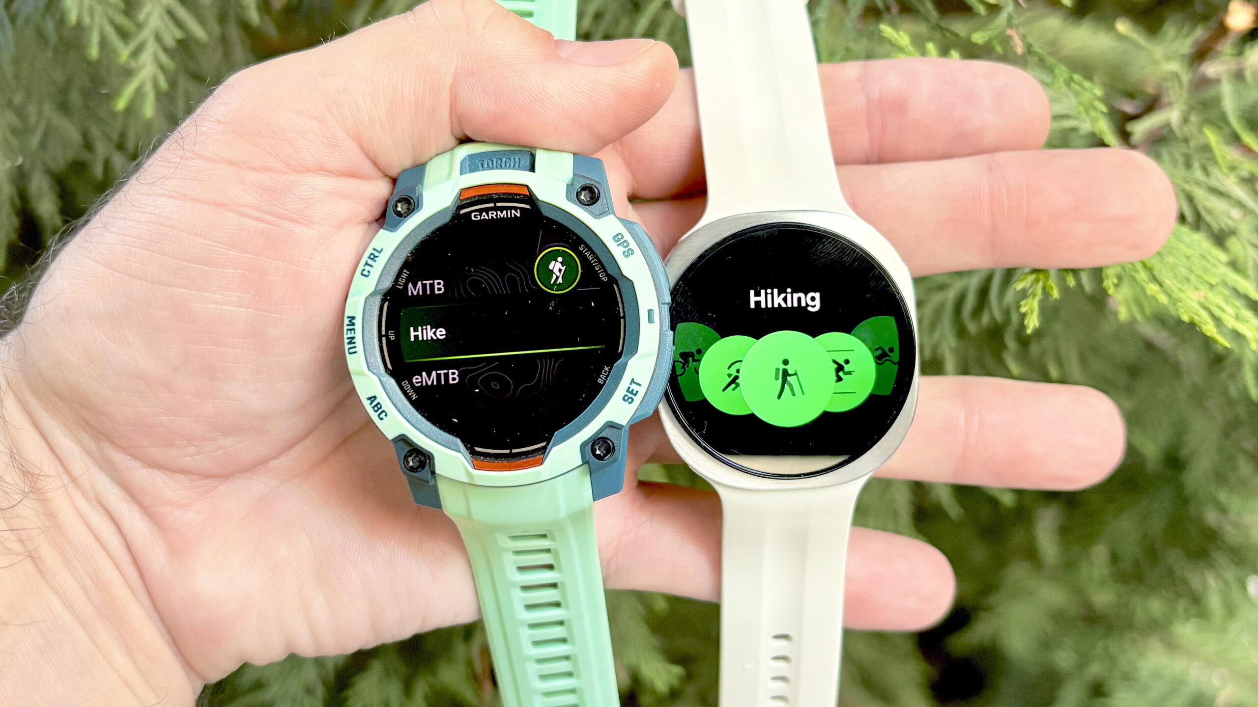 لقطة مقربة لساعة Garmin Instinct 3 بجانب Samsung Galaxy Watch 9 مع أشجار دائمة الخضرة في الخلفية