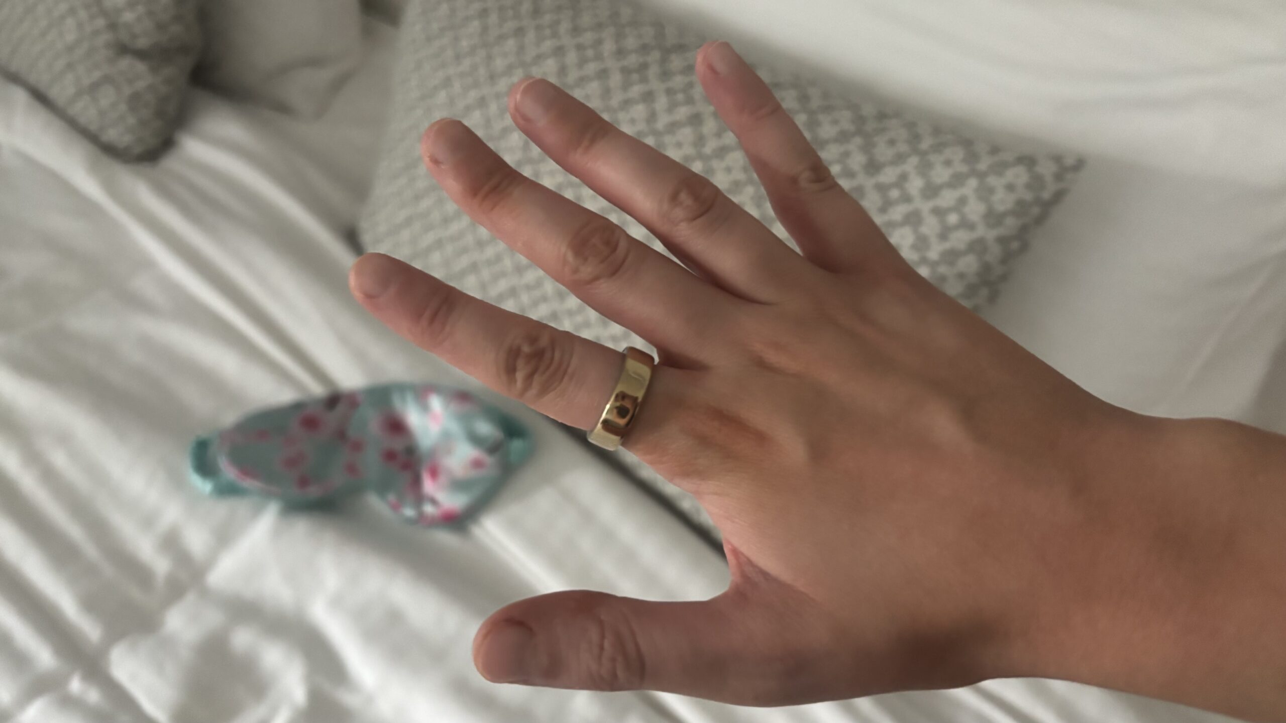 يد امرأة تعرض خاتم Oura Ring 5 أمام سرير فندق مرتب حديثًا.